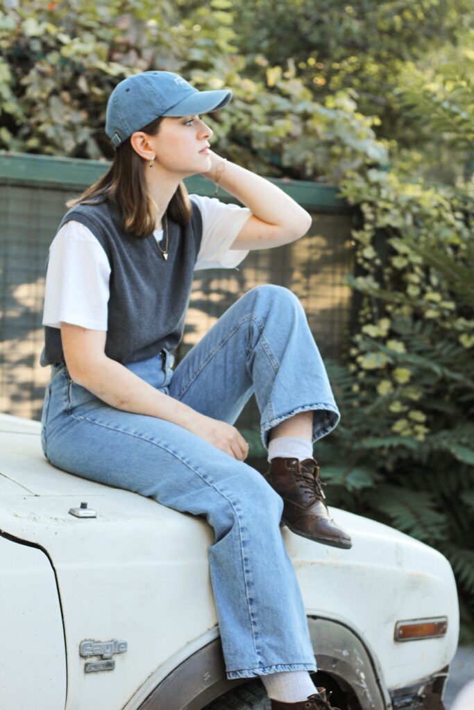 Young woman in casual attire sitting on a vintage car hood outdoors.