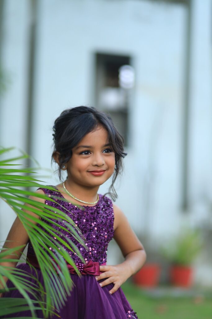 Delightful outdoor portrait of a smiling young girl in a purple dress amidst greenery.