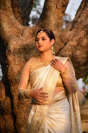 A woman in a golden saree stands gracefully by a tree, exuding cultural elegance.