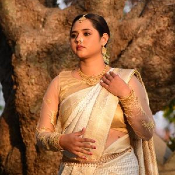 A woman in a golden saree stands gracefully by a tree, exuding cultural elegance.